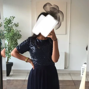 Mörkblå glittrig långklänning - Snygg mörkblå långklänning med glittrig paljett-topp och rund halsringning. Kjolen är i ett mjukt, flowy tyg och klänningen har korta ärmar. Perfekt för dig som vill sticka ut med lite extra glitter.