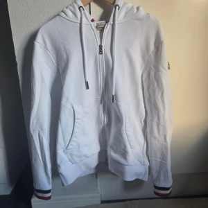 Vit hoodie från Moncler med dragkedja - Vit hoodie från Moncler med huva och snörning, dragkedja framtill och två fickor. Ribbade muddar med röd, blå och vit rand på ärmslut och huva. Moncler-logga på vänster ärm. Tillverkad i mjuk bomull, perfekt för chill dagar.