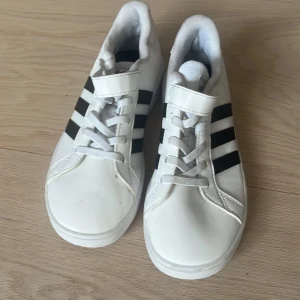 Vita Adidas sneakers med svarta ränder - Snygga vita original  oanvända Adidas sneakers med klassiska svarta ränder på sidorna. Skorna har snörning och kardborreband upptill för extra smidig passform. Tillverkade i syntetmaterial med platt sula och rund tå. Perfekta för en sportig och stilren look. Storlek 33.