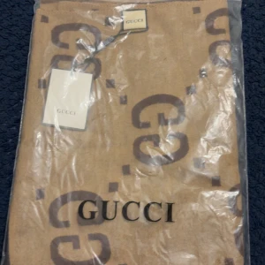 Beige halsduk med GG-logga från Gucci - Snygg beige halsduk från Gucci med stora GG-loggor i mörkbrunt över hela tyget. Halsduken har en rektangulär form och är tillverkad i ett mjukt material som känns lyxigt. Perfekt för dig som vill ha en klassisk designeraccessoar med tydlig branding.
