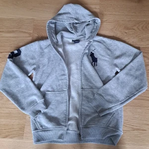 Grå hoodie från Ralph Lauren - Snygg grå hoodie från Ralph Lauren med dragkedja och stor broderad marinblå logga på bröstet. Tröjan har huva, två fickor fram och mjukt innerfoder. DRAGKEDJAN FUNGERAR INTE! Men annars inga defekter!