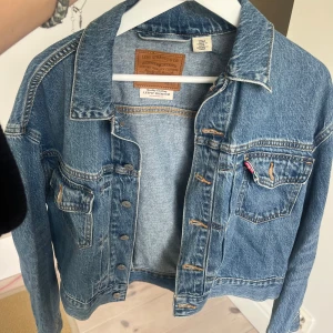 Levi's jeansjacka i klassisk blå denim - Säljer en klassisk blå jeansjacka från Levi's Premium med snygga sömmar, bröstfickor med knappar och ikonisk röd Levi's-tagg. Jackan har en rak passform och är tillverkad i slitstarkt bomullsjeans. Perfekt för dig som vill ha en tidlös och cool look.