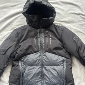 Svart och grå dunjacka Sail Racing - Säljer en svart och grå dunjacka från Sail Racing med huva och flera dragkedjeförsedda fickor. Jackan har quiltad design på framsidan och är tillverkad i polyester och dun. Perfekt för kalla dagar och har snygga detaljer som logga på bröstet och ärmen.