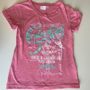 Rosa Costa Azul t-shirt från Mywear - Snygg rosa t-shirt från Mywear Young med Costa Azul-tryck i turkos och vitt framtill. T-shirten har rund hals, korta ärmar och är gjord i mjuk bomull och polyester. Perfekt för dig som gillar surf vibes och tropiska motiv.