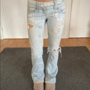Ljusblå slitna lågmidjade jeans - Säljer ett par ljusblå jeans med bootcut passform och flera slitningar och hål för en riktigt edgy look. Jeansen har låg midja och klassisk femficksdesign. Perfekta för dig som gillar en avslappnad och trendig stil. Midja 37 innerben 79