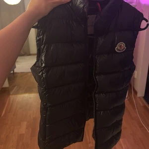 Svart dunväst från Moncler - Snygg svart dunväst från Moncler med quiltad design och klassisk logotyp på bröstet. Västen har hög krage och dragkedja framtill. Perfekt att slänga över en hoodie för extra värme. Materialet är glansigt och känns lyxigt. 