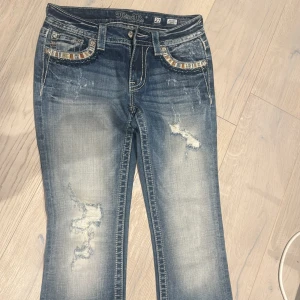 Miss Me blå bootcut jeans med paljettdetaljer - Snygga blå jeans från Miss Me med bootcut passform. Jeansen har slitna detaljer framtill och coola paljetter och broderier på fickorna både fram och bak. Tydliga kontrastsömmar. Perfekta för dig som gillar statement-jeans! Aldrig använda eftersom de är för små. De är i strl 27 men skulle säga att de är mer som en 26. Skicket är som nytt 