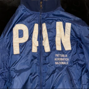 Blå vindjacka Aeronautica Militare - Snygg blå vindjacka från Aeronautica Militare med stora vita bokstäver och broderade flaggor på ryggen. Jackan har dragkedja, ribbade muddar och meshfoder. Texten 'Pattuglia Acrobatica Nazionale' framtill ger en unik touch.