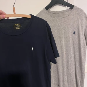 2-pack Polo Ralph Lauren t-shirts - Två klassiska t-shirts från Polo Ralph Lauren, en i marinblått och en i ljusgrått. Båda har rund hals, korta ärmar och den ikoniska broderade loggan på bröstet. Tillverkade i mjuk bomull för skön komfort och stilren look. Den blå är i storlek L men den är i en modell som gör den en M.