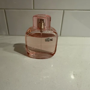 Rosa Lacoste parfymflaska - Snygg rosa parfym från Lacoste med modern design och ikonisk krokodillogo. Perfekt för dig som vill ha en stilren accessoar på hyllan. 