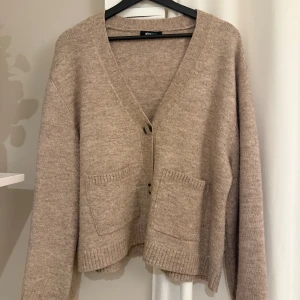 Beige kofta från Gina Tricot - Mysig beige kofta från Gina Tricot med v-ringning, två stora fickor framtill och knappar. Koftan är stickad och har långa ärmar samt ribbade detaljer vid ärmslut och nederkant. Perfekt för lager-på-lager-stil.