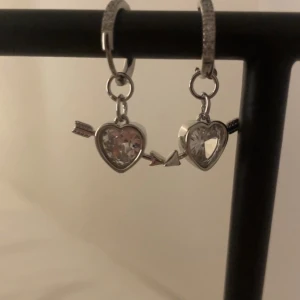 Silverörhängen med hjärtan och pil vit - Ett par snygga örhängen i silverfärg med glittrande hjärtformade hängen och pil-detaljer. Hängena har en kristall-liknande sten i mitten som ger extra bling. Perfekta för dig som gillar romantiska och trendiga accessoarer. Rostfri 