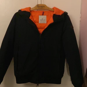 Svart Moncler pufferjacka med orange foder - Säljer en svart pufferjacka från Moncler med orange quiltat foder och huva. Jackan har dragkedja framtill och ribbade muddar. Klassisk Moncler-logga på ärmen och snygg kontrast mellan det svarta yttermaterialet och det orange fodret. Perfekt för kalla dagar.