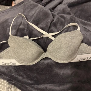 Grå Calvin Klein bh 75B - Snygg grå bh från Calvin Klein med vit elastisk kant och logga. Mjuk och bekväm med justerbara axelband och lätt vadderade kupor.