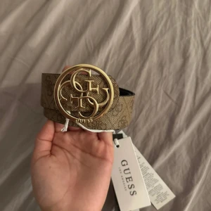 Guess bälte med guldspänne och logomönster - Snyggt bälte från Guess med stort, rundat guldspänne och GG-logga. Bältet har ett brunt och svart logomönster på båda sidor och är tillverkat i syntetmaterial. Perfekt för att lyfta din outfit med en exklusiv touch och tydlig branding. Den är aldrig använd och har kvar lapparna. Den kommer i en guess påse som tillhör den. Byst 85-90cm, midja 72-79cm, höfter 93-98cm