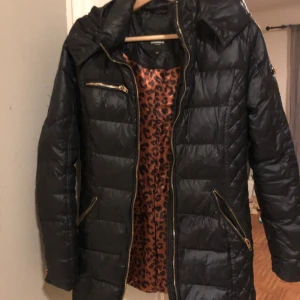 Svart dunjacka Rockandblue med leopardfoder - Svart dunjacka från Rockandblue med snygga guldfärgade dragkedjor och coolt leopardmönstrat foder. Jackan har quiltade partier, hög krage och flera fickor med dragkedja. Perfekt för dig som vill ha en jacka med attityd och detaljer som sticker ut.