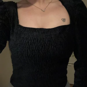 Svart långärmad blus med puffärm - Säljer en svart blus med lång puffärm och fyrkantig urringning. Toppen har en smockad, rynkad struktur som ger en cool och trendig look. Perfekt för dig som gillar att sticka ut med stilrena detaljer och markerade axlar.