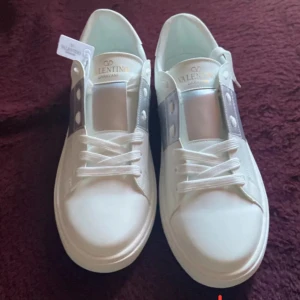 Valentino Garavani vita sneakers - Säljer ett par fräscha vita Valentino Garavani sneakers med silvrig detalj över vristen och klassisk snörning. Skorna är i skinn och har en chunky sula med mönster undertill samt nitar baktill för extra edge. Perfekta för dig som vill sticka ut med stil.