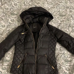 Svart pufferjacka från Rockandblue - Säljer en svart pufferjacka från Rockandblue med snygga guldfärgade dragkedjor och quiltade partier på sidorna. Jackan har huva och flera praktiska fickor med dragkedja. Perfekt för kyliga dagar och riktigt stilren look.