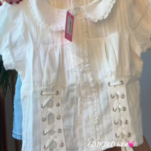 Vit blus med snörning från Edikted - Supersöt vit blus från Edikted med korta puffärmar, spetsdetaljer vid kragen och knappar framtill. Blusen är croppad och har snygg snörning i sidorna som ger en unik look. Perfekt för dig som gillar romantisk och trendig stil.