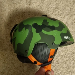 Grön camo hjälm från WARP - Säljer en grym hjälm från WARP i grönt camouflage-mönster med svarta och mörkgröna detaljer. Hjälmen har orange hakrem och ventiler på sidorna. Perfekt för dig som vill sticka ut och samtidigt vara säker. Passar huvudstorlek 54/55.