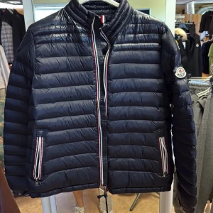 Mörkblå dunjacka från Moncler - Säljer en mörkblå dunjacka från Moncler med klassisk quiltad design och vita, röda och blå detaljer vid dragkedja och fickor. Jackan har ståkrage, Moncler-logga på ärmen och är tillverkad i glansig polyester. Perfekt för kyliga dagar.