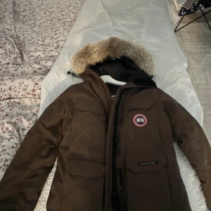 Brun Canada Goose parkas XS - Säljer en brun Canada Goose parkas med fluffig päls på huvudet och klassisk logga på bröstet. Jackan har stora fickor framtill, dragkedja och knappar. Perfekt för kalla vinterdagar och riktigt snygg.