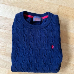 Mörkblå kabelstickad tröja Morris S - Kabelstickad mörkblå tröja från Morris i 100% merinoull. Klassisk rund halsringning, långärmad och med röd broderad logga på bröstet. Perfekt för dig som gillar stilrena och tidlösa plagg med snygga detaljer. Storleken på tröjan är L men den sitter som S. Skicket är galet bra och om du har några funderingar är det bara att höra av sig