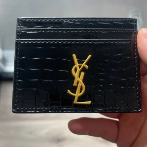 Svart kortfodral från Saint Laurent - Säljer ett svart kortfodral från Saint Laurent med krokopräglat mönster och guldig YSL-logga framtill. Perfekt för att hålla koll på dina kort med stil. Gjord i glansigt skinn och har flera kortfack.