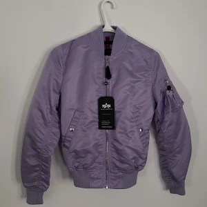 Bomberjacka från Alpha Industries - Snygg lila bomberjacka från Alpha Industries med klassisk passform, ribbade muddar och silverfärgad dragkedja. Jackan har fickor med tryckknappar och en ikonisk ärmficka med zip och tag. Tillverkad i glansig polyester som ger en modern vibe.
