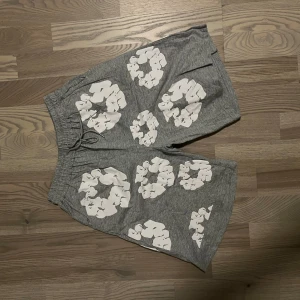 Grå shorts med vit blommig print - Säljer ett par grå shorts med coola vita blommiga tryck över hela. De har elastisk midja med snörning för skön passform och är gjorda i mjukt bomullsmaterial. Perfekta för chill dagar eller när du vill sticka ut lite extra.
