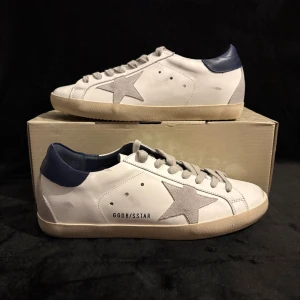 Golden Goose Superstar sneakers vit/blå - Golden Goose Superstar sneakers i vitt skinn med blå häl och grå mockadetaljer. Snygg stjärna i grått på sidan, beige platt sula och klassisk snörning. Insidan är märkt med 'SUPER-STAR' och 'Genuine Leather'. Perfekt för dig som gillar italiensk design och streetstyle.