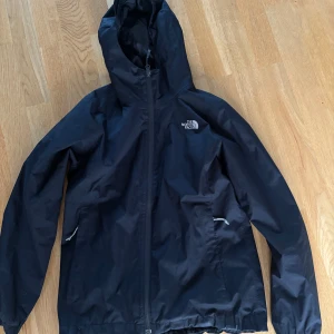 Svart regnjacka från The North Face - Svart regnjacka från The North Face med huva och hel dragkedja framtill. Jackan har två fickor med dragkedja och meshfoder på insidan. Perfekt för blöta dagar och enkel att slänga på sig när det regnar. Säljer den då den har blivit för liten för mig. 
