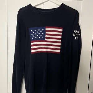 Mörkblå stickad tröja GANT med USA-flagga - Snygg mörkblå stickad tröja från GANT i storlek M. Framtill finns en stor broderad USA-flagga i blått, vitt och rött. På ena ärmen står GANT NHCT 1949 broderat i vitt. Tröjan har rund hals och långa ärmar, perfekt för dig som gillar preppy stil.