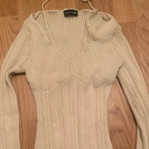 Beige ribbad stickad tröja PrettyLittleThing - Snygg beige stickad tröja från PrettyLittleThing med ribbad struktur och v-ringad hals. Tröjan har långa ärmar och en figurnära passform som framhäver siluetten. Perfekt för dig som gillar stilrena och trendiga plagg med lite edge.