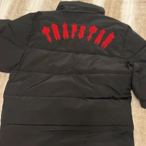Trapstar svart pufferjacka med röd logga - Svart pufferjacka från Trapstar med fet röd logga på ryggen och rund patch på bröstet. Jackan har hög krage, dragkedja med unik T-detalj och två fickor framtill. Perfekt för kalla dagar och riktigt street i stilen.