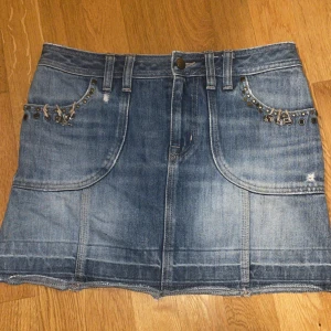 Y2K vintage kjol från Karen Millen - Exklusiv jeanskjol från Karen Millen, äkta 2000-talsmodell i denim med fickor fram och bak samt detaljer i metall och små charms. Klassisk Y2K-stil som påminner om tidiga 00-talskollektioner. 