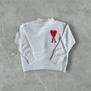 Vit stickad tröja från Ami Paris(fast pris) - Vit stickad tröja från Ami Paris med röd hjärtlogga och bokstaven A på bröstet. Tröjan har ribbade muddar, hög hals och är långärmad. Perfekt för dig som gillar stilrena och trendiga plagg med en twist.
