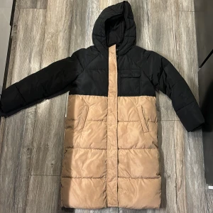 Lång vinter eller höst jacka  - Svart och beige dunjacka längre modell. Hel och fin, men någon liten skönhetsfläck syns. Har väldigt mycket mer att ge. Strlk 146.