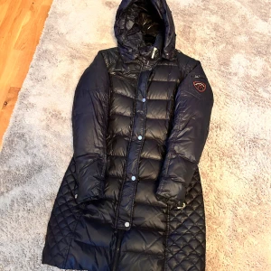 Svart lång dunjacka med huva - Säljer en svart lång dunjacka från Rockandblue med huva och quiltade detaljer. Jackan har dragkedja och knappar framtill, samt en patch på ärmen. Perfekt för kalla dagar och har en snygg, figurnära passform.