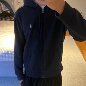 Mörkblå hoodie från Polo Ralph Lauren - Snygg mörkblå hoodie från Polo Ralph Lauren med dragkedja och klassisk vit logga på bröstet. Tröjan har huva med snörning och två fickor framtill. Perfekt för en chill och stilren look. Modellen är 186, 70 kg