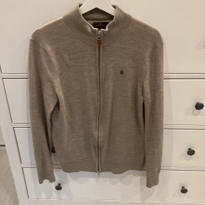 Beige kofta i merinoull från Morris - Säljer en stilren beige kofta från Morris i 100% merinoull. Koftan har hel dragkedja framtill, ribbad krage och långa ärmar. Perfekt för lager-på-lager och snygg till både jeans och chinos. Klassisk Morris-logga broderad på bröstet.