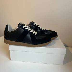 Svarta Maison Margiela med mockadetaljer - Maison Margiela sneakers i svart skinn och mocka med kontrasterande vita snören och beige gummisula. Klassisk rund tå och platt sula, stilren design med diskreta paneler och mjuk insida. Perfekt för dig som gillar minimalistisk och clean look. Hör av er vid någon fråga eller fundering. Priset kan diskuteras vid snabb affär!✅