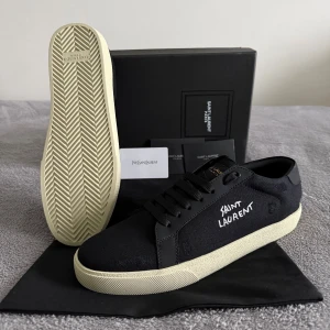  Saint Laurent skor - Svarta sneakers från Saint Laurent med vit broderad logga på sidan. Snygg kontrast mellan svart canvas och krämfärgad platt gummisula. Klassisk låg modell med rund tå och svarta snören. Perfekt för dig som gillar stilrena och exklusiva sneakers. 