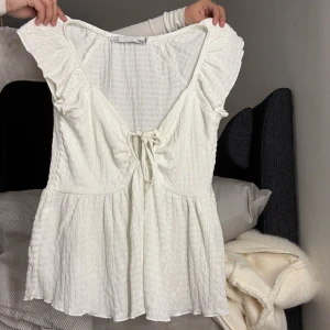 Vit blus med volang och knyt - Supersöt vit blus från 157 med volangärmar och v-ringning. Toppen har knyt framtill och är tillverkad i ett lätt, luftigt bomullstyg med struktur. Perfekt för en fräsch och somrig look med flowig passform och romantiska detaljer.