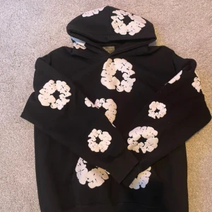 svart denim tears hoodie - Svart hoodie från Denim tears med stora vita blommönster över hela tröjan och huva. Oversized fit och mjukt bomullsmaterial, perfekt för dig som vill sticka ut med en unik streetstyle-look.