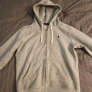Grå hoodie från Polo Ralph Lauren - Snygg grå hoodie från Polo Ralph Lauren med dragkedja, huva med snörning och två fickor framtill. Klassisk logga broderad på bröstet. Perfekt för chill dagar och enkel att matcha med jeans eller joggers. Mjuk och skön bomullsmix som håller dig varm.