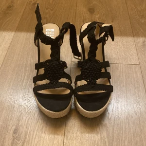 Svarta sandaler med flätad plattform - Snygga svarta sandaler från Bullboxer med flätade remmar och plattformssula i rep-look. Remmarna är grovt flätade och har spänne vid ankeln. Perfekt för dig som gillar boho-vibe och vill ha lite extra höjd. Sulan är beige och ger en cool kontrast mot de svarta detaljerna.