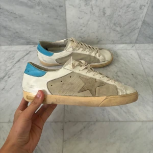 Golden Goose vita sneakers med blå detalj - Säljer ett par snygga Golden Goose sneakers i vitt och beige med blå häl och klassisk stjärndetalj på sidan. Skorna har snörning, platt sula och rund tå. Materialmix av skinn och mocka ger en cool och avslappnad vibe. Perfekt för dig som gillar streetstyle.