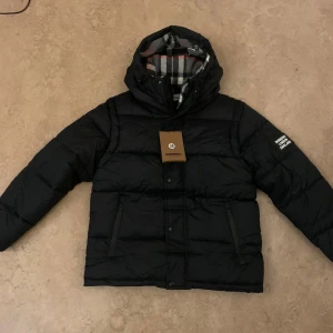 Svart pufferjacka från Burberry - Svart pufferjacka från Burberry med huva och två dragkedjefickor framtill. Jackan har ett stort Burberry London England-märke på ärmen och knappar med Burberry-logga. Insidan har rutigt foder i grått, vitt och rött. Perfekt för kalla dagar.
